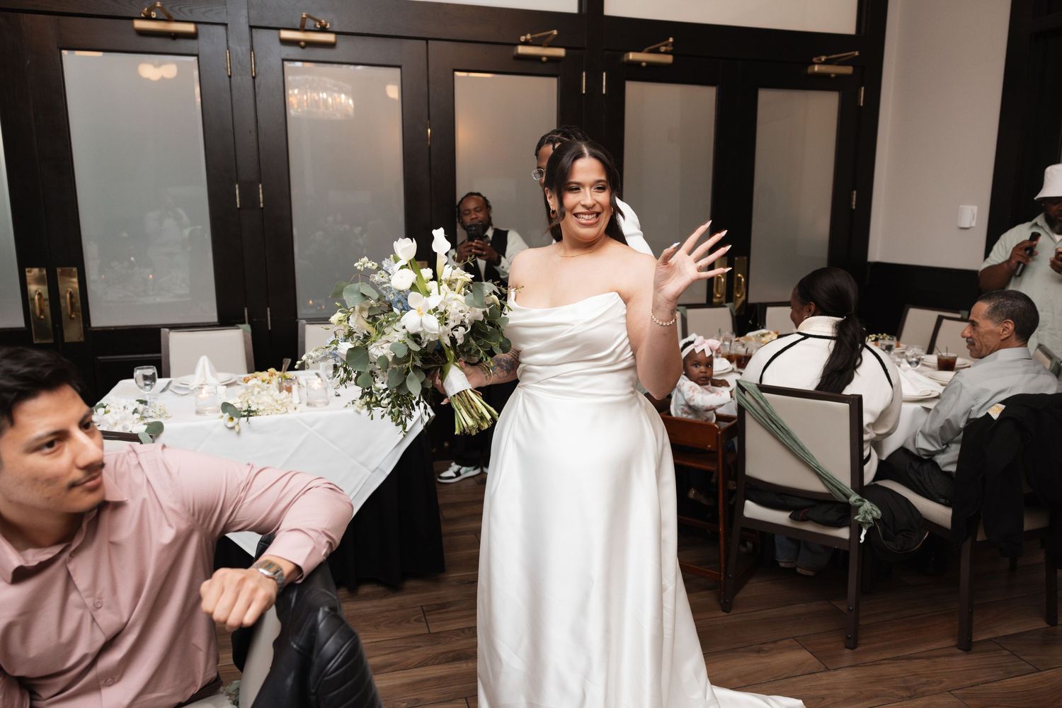 Bride in strapless white gown holding floral bouquet greeting guests at wedding reception.
