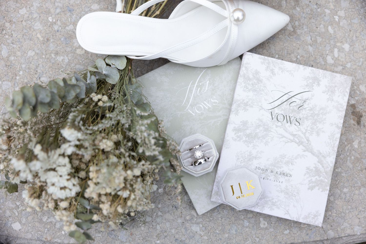 Elegant wedding flat lay featuring white heels, dried floral bouquet, diamond rings, and a grey FOWN wedding program on stone.