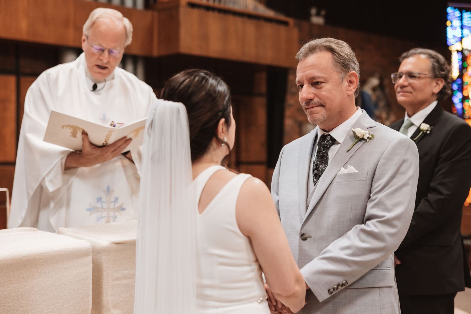 Bride and groom holding hands at church wedding ceremony while officiant reads from book at altar.