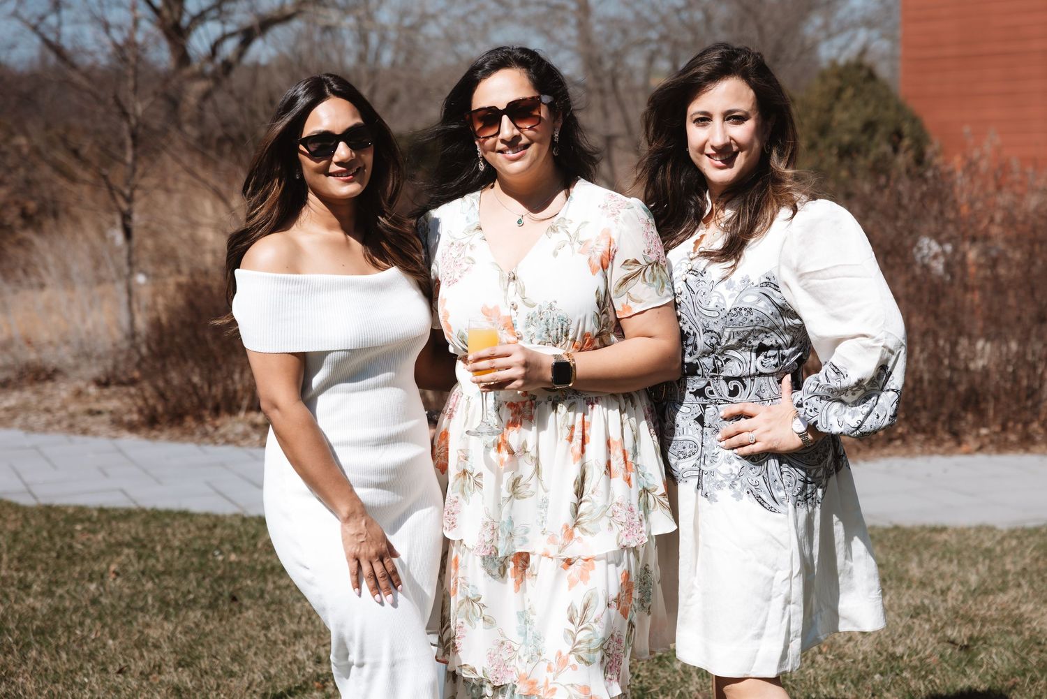 Three women in elegant white and floral dresses posing outdoors on a sunny day at a celebration.