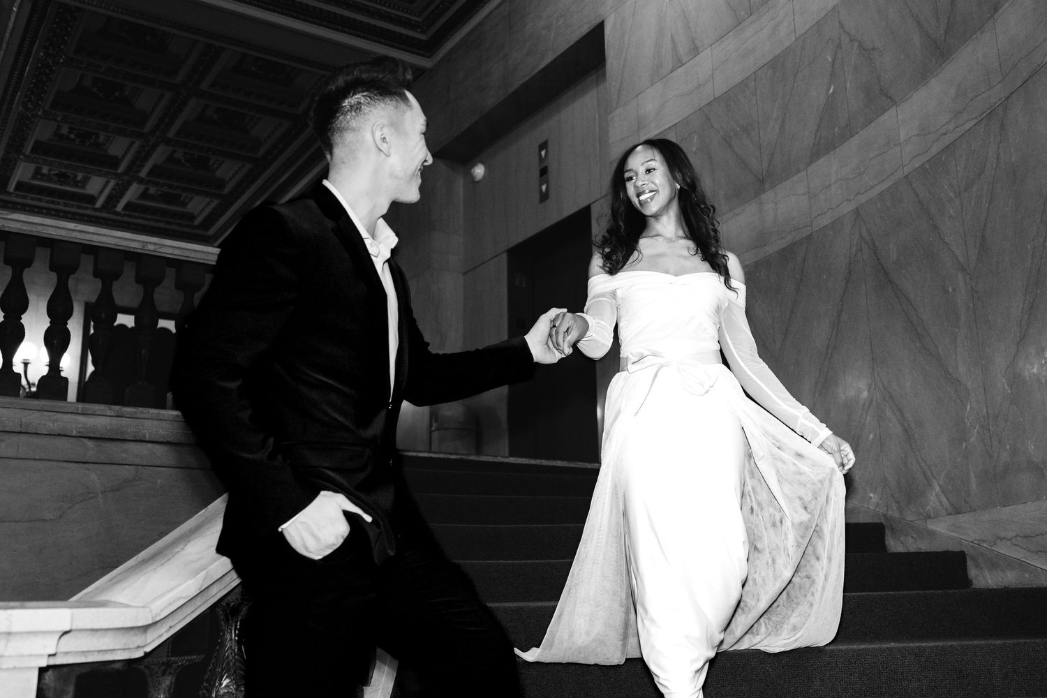 Black and white photo of groom holding brides hand as she lifts gown on staircase.