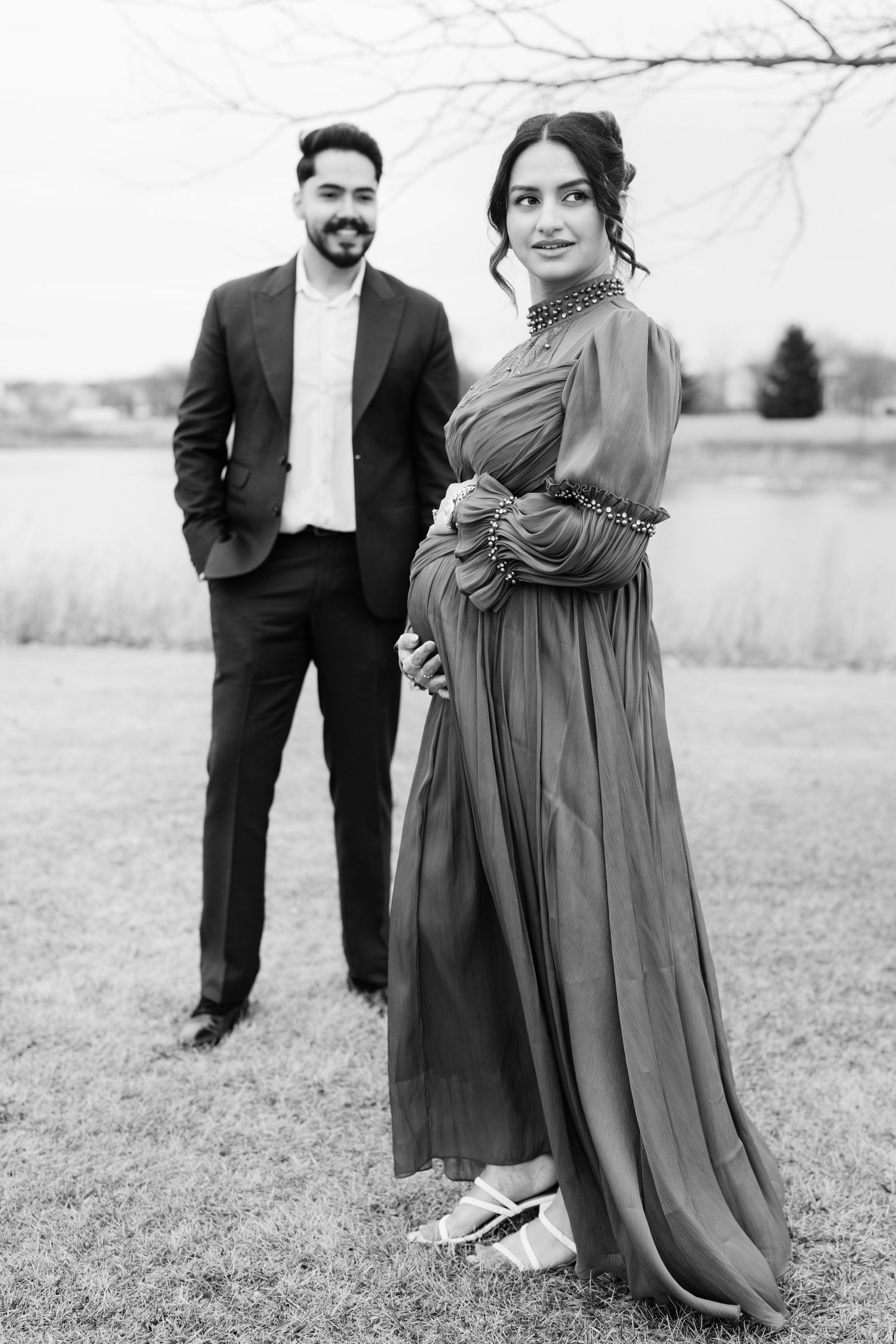 Black and white maternity photo of couple posing outdoors near a lake, woman in elegant gown.