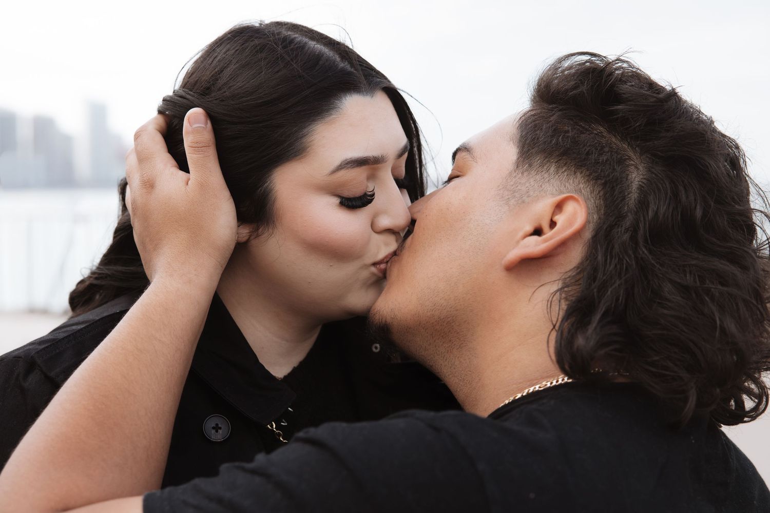 Couple shares romantic kiss outdoors with soft waterfront background in urban setting.