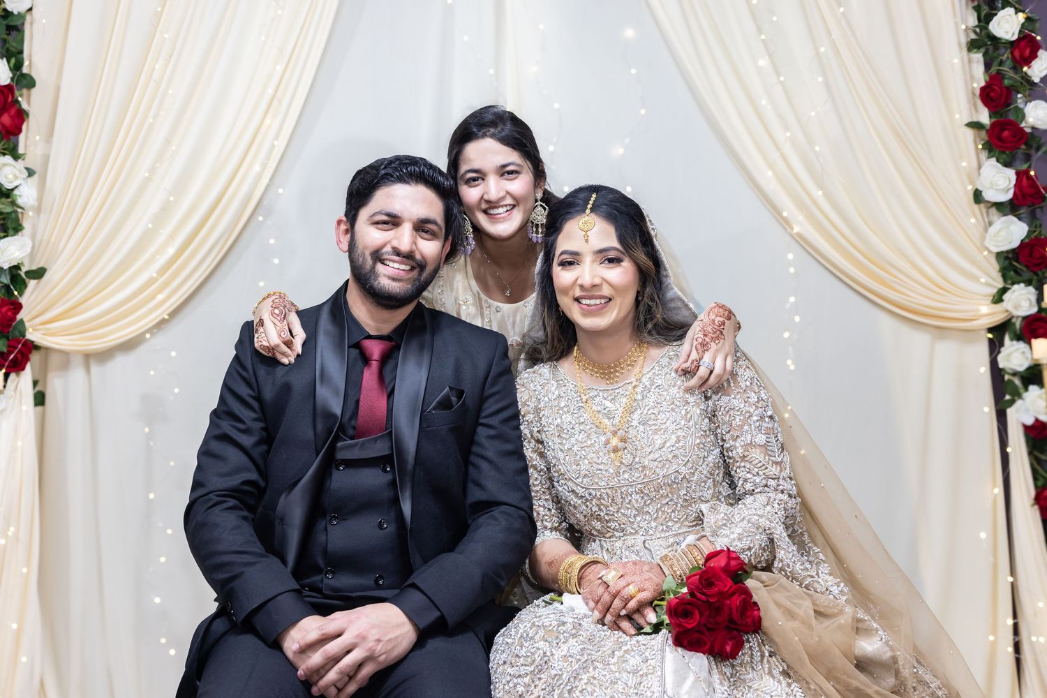 Bride in gold bridal lehenga sits with groom in black suit and smiling woman at floral wedding backdrop.