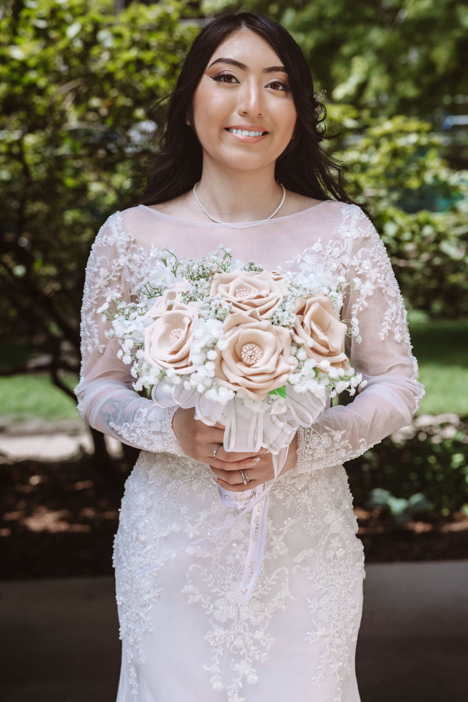 Smiling bride in white lace long sleeve gown holding blush rose bouquet outdoors.