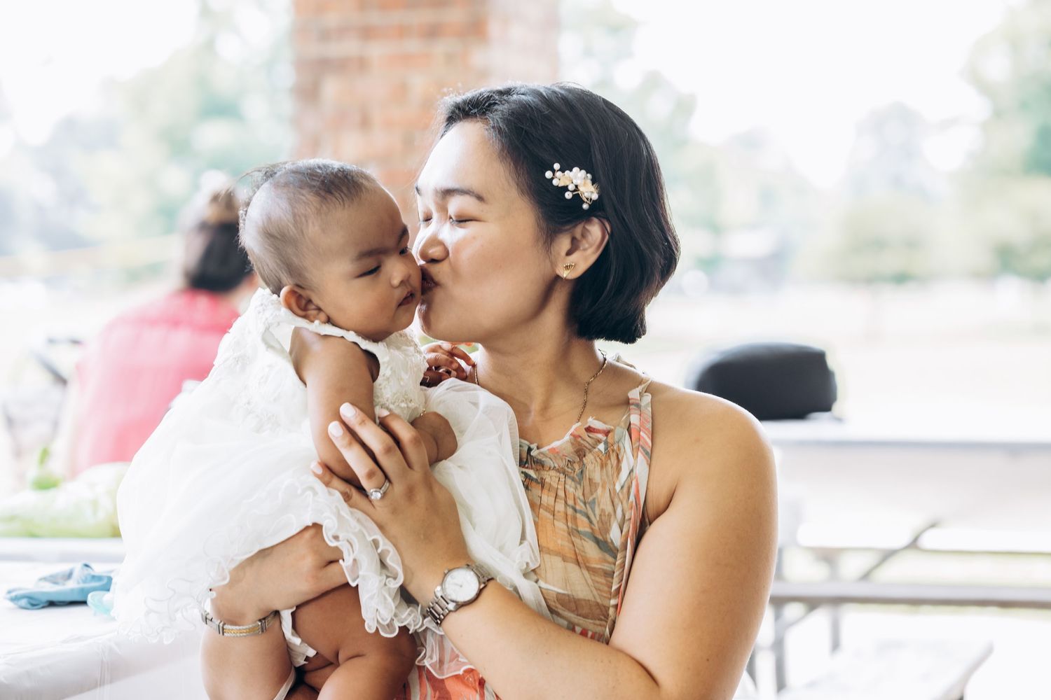 Loving mother in floral dress kisses baby in white baptism gown outdoors.