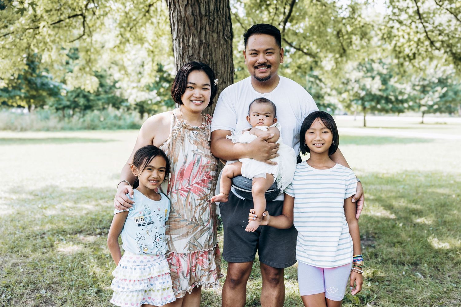 Happy Asian family of five poses together under tree in summer park.
