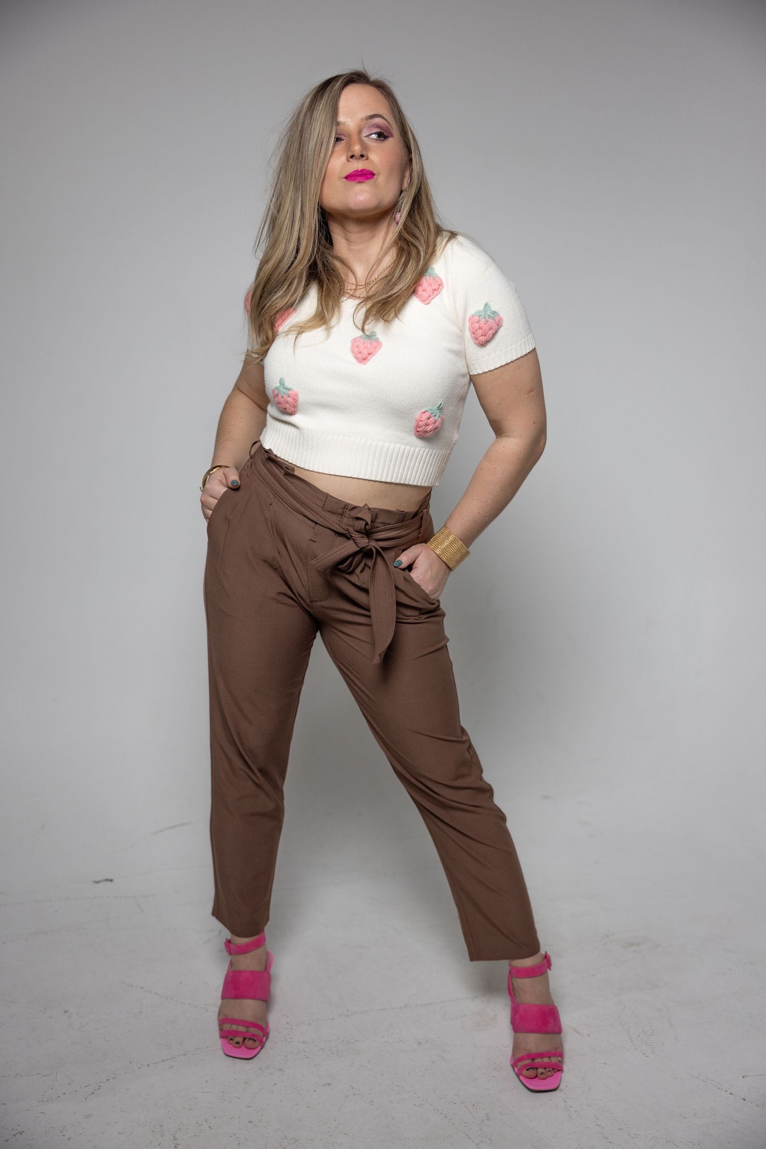 Woman posing in a white strawberry crop top, brown tie-waist pants, and pink heels in a studio.