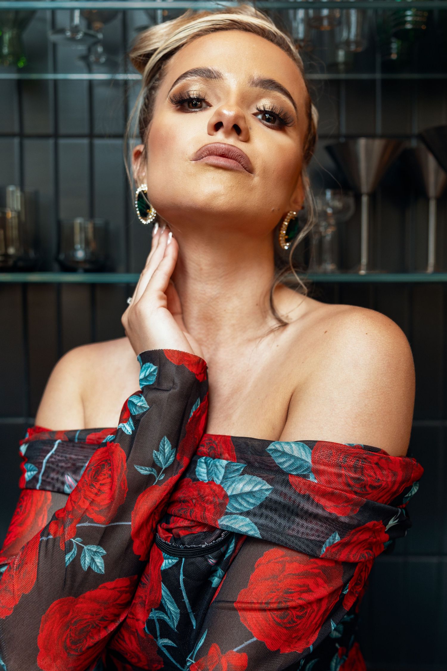 Close-up of woman in off-shoulder floral dress with bold makeup touching her neck.