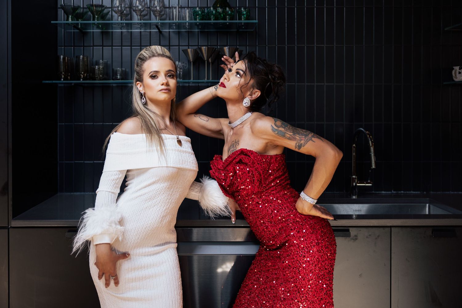 Two women in white feathered and red sequin gowns posing in dark modern kitchen.