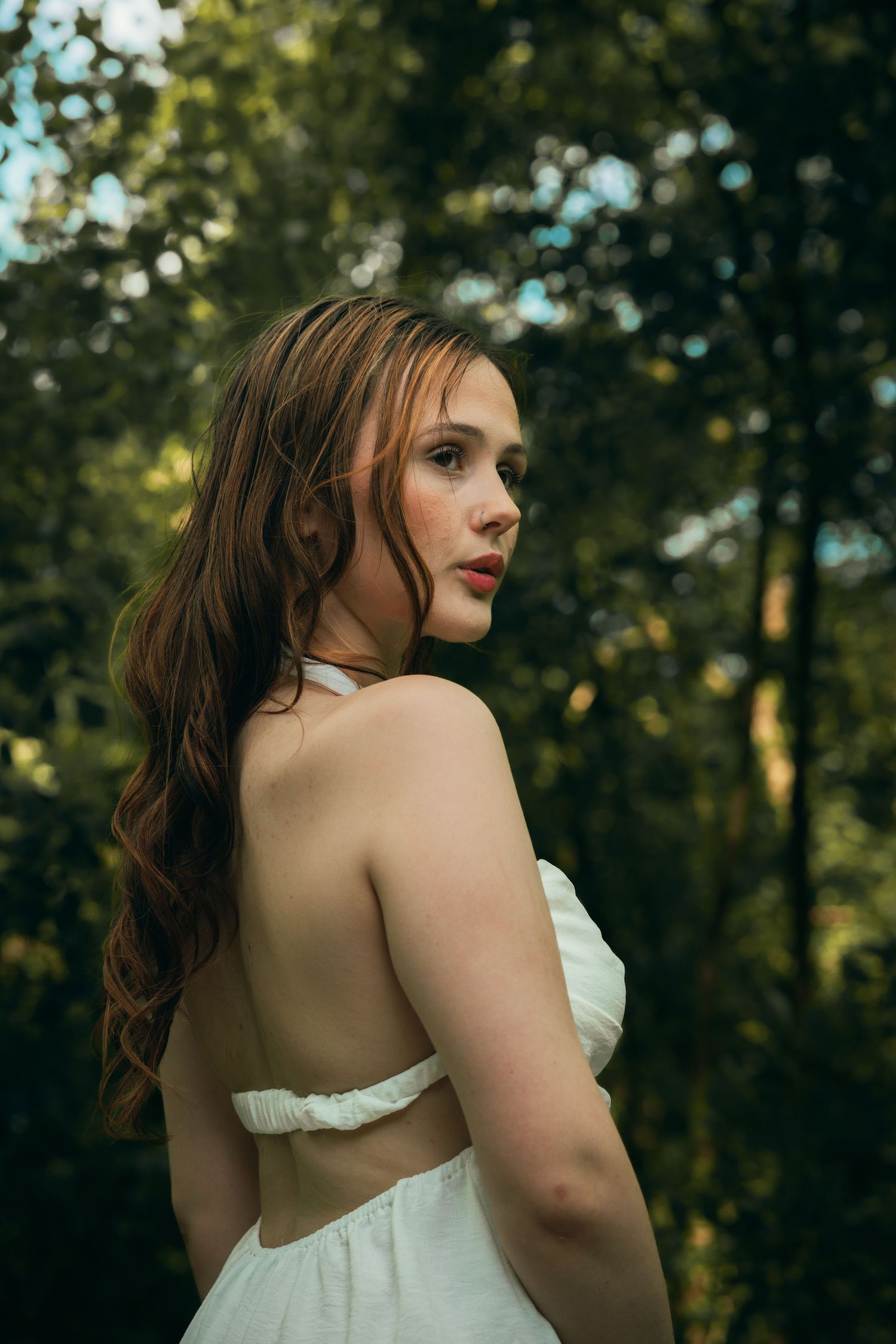 A young woman with auburn hair in a backless white dress looks over her shoulder in a sunlit forest.