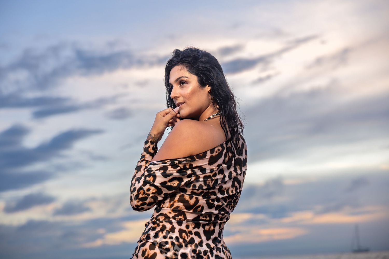 Woman in leopard print dress posing against colorful cloudy sky at beach.