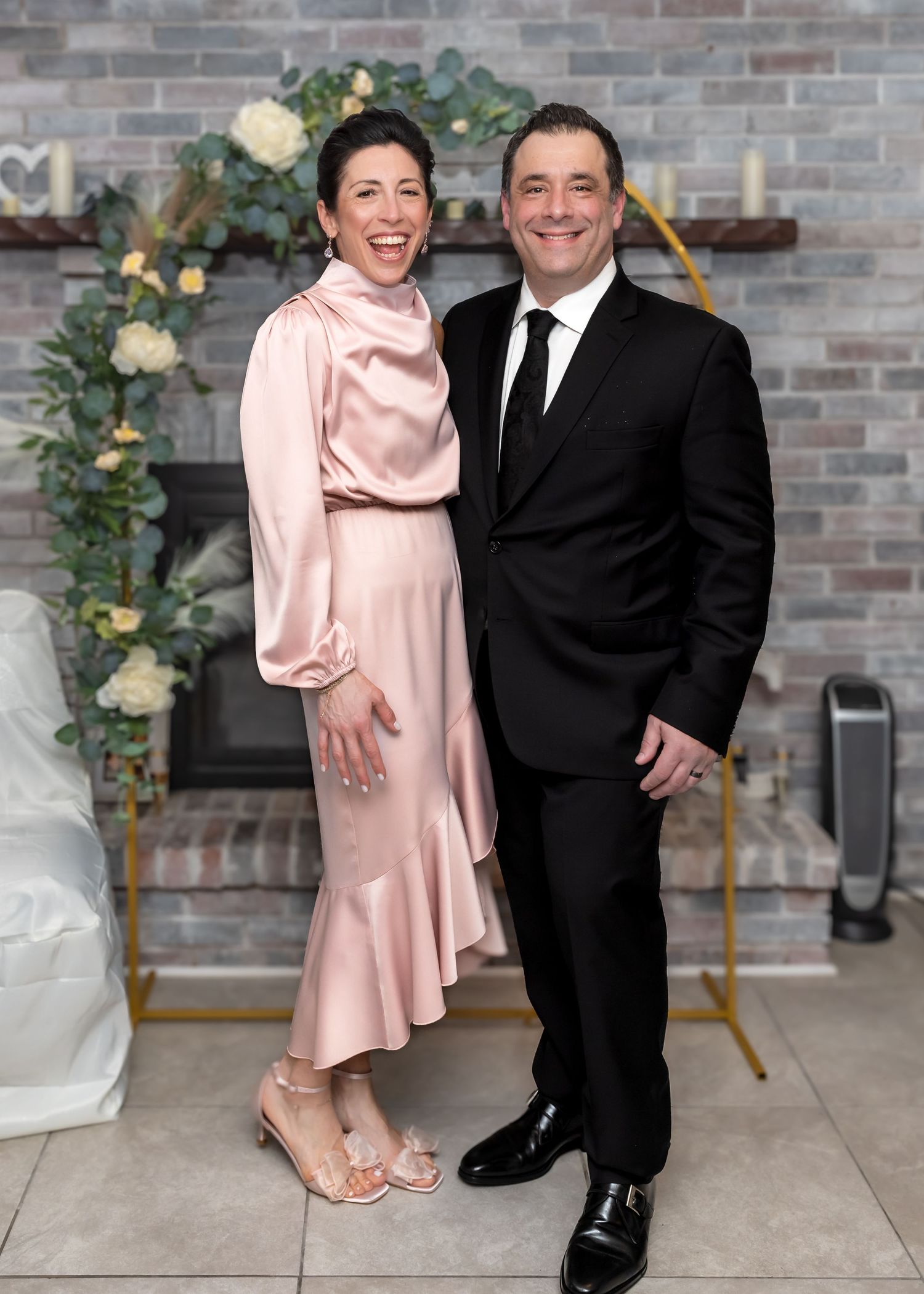 Smiling couple in formal attire standing before a gold floral arch at a wedding celebration.
