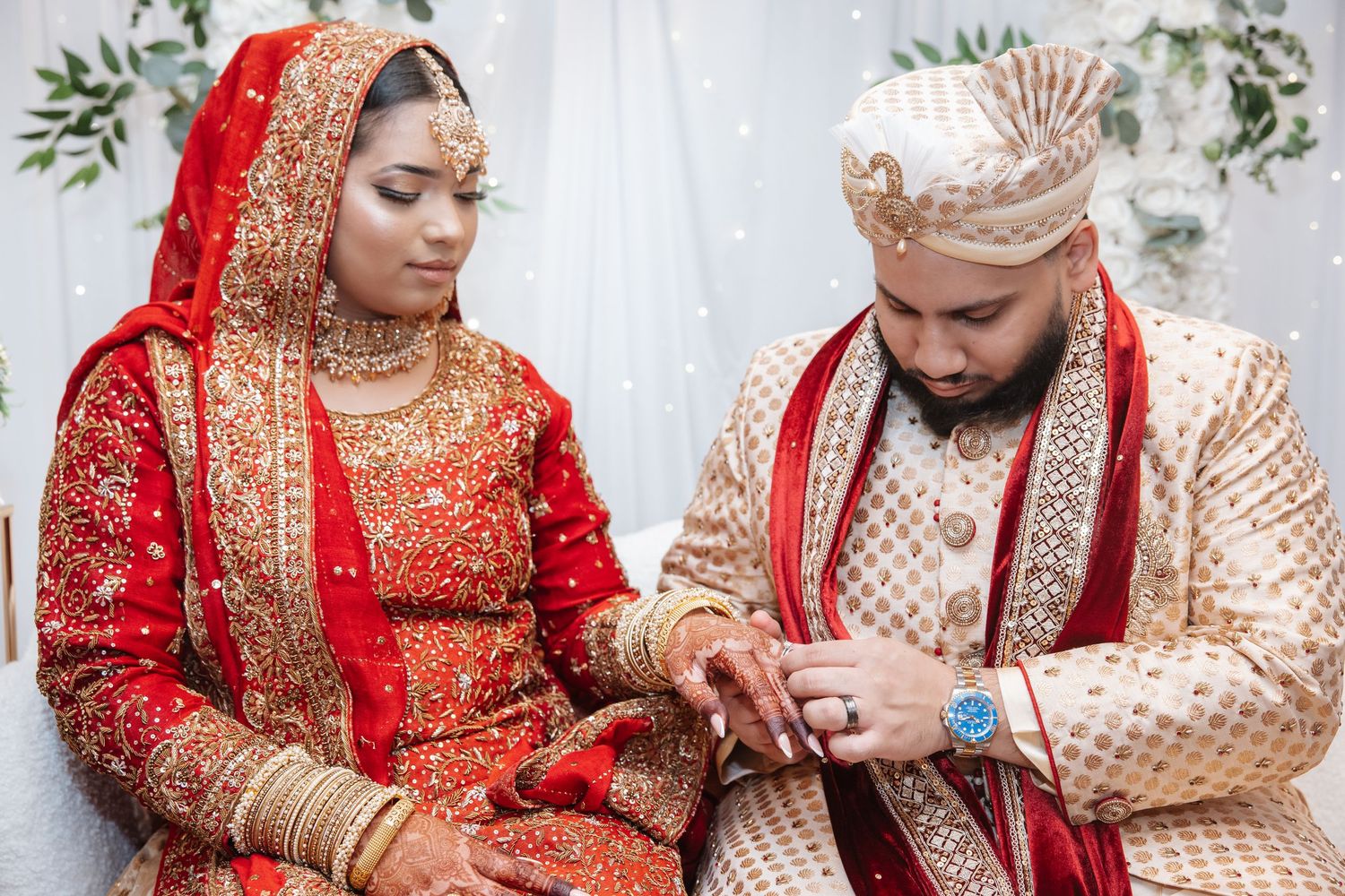 Indian bride and groom in traditional red and gold wedding attire during ring exchange ceremony.