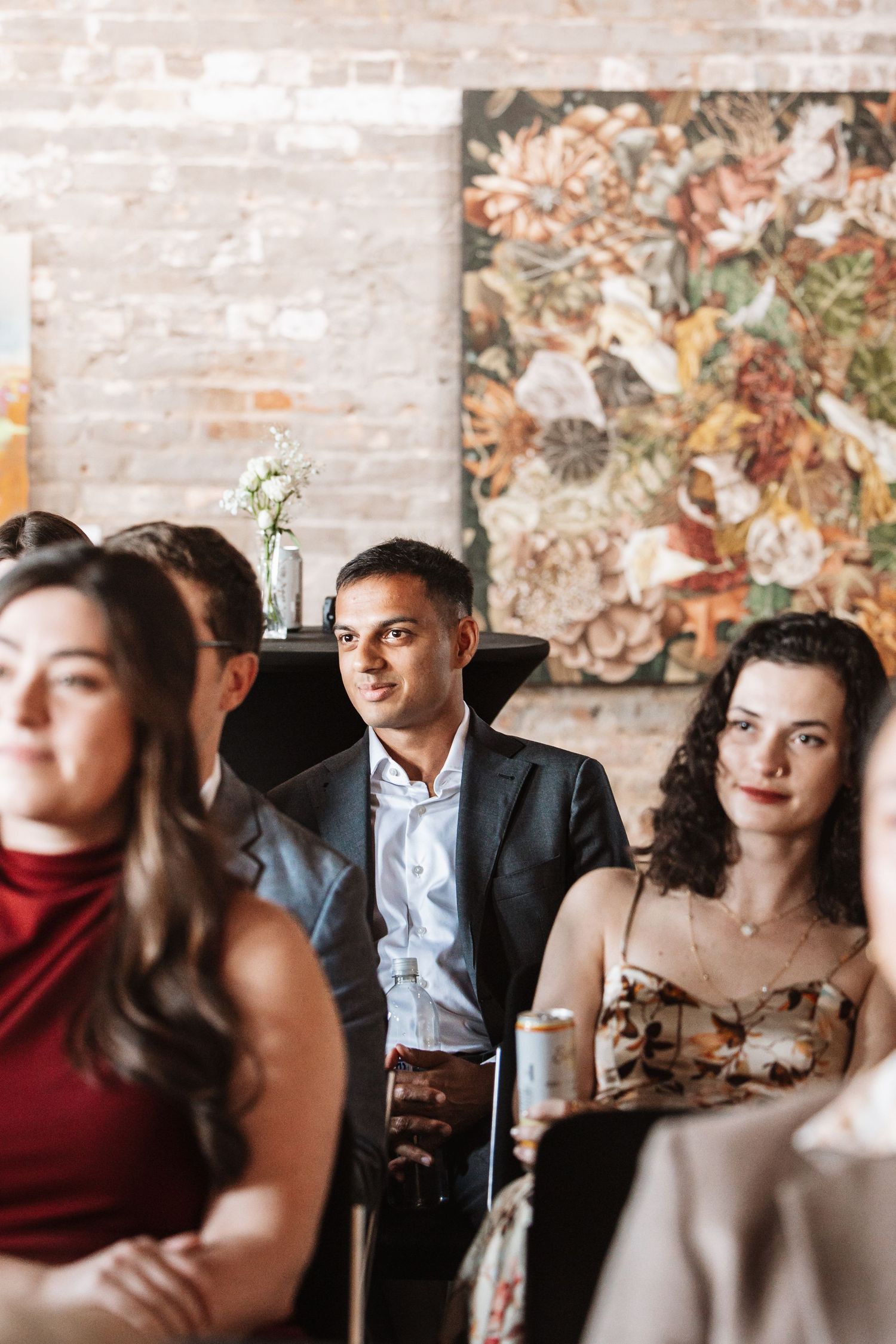 Guests seated at a formal event with floral artwork on brick wall in background.