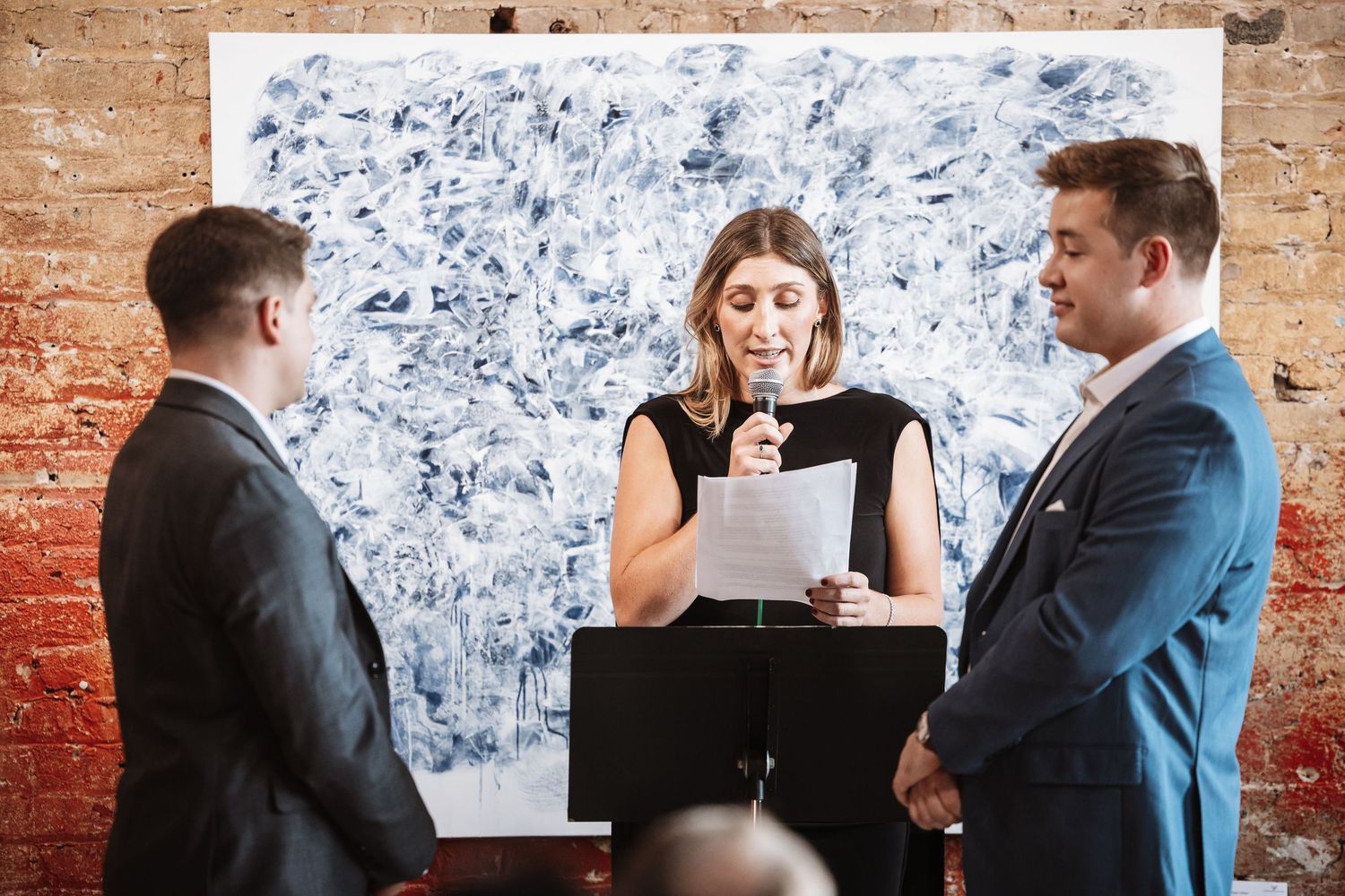 Female officiant reads from papers as two grooms stand attentively during their art gallery wedding.