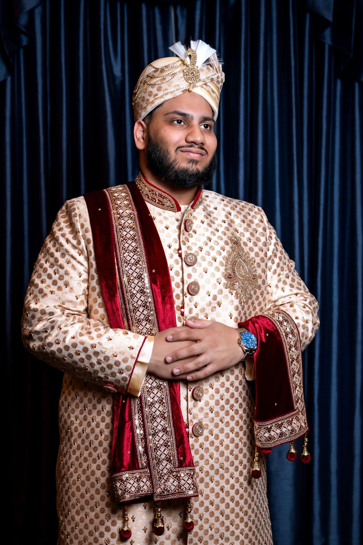 Groom in gold embroidered sherwani with red velvet stole and turban smiling at South Asian wedding ceremony.