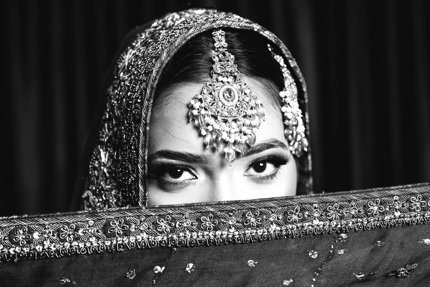 Black and white portrait of an Indian bride in traditional jewelry and veil covering her face.