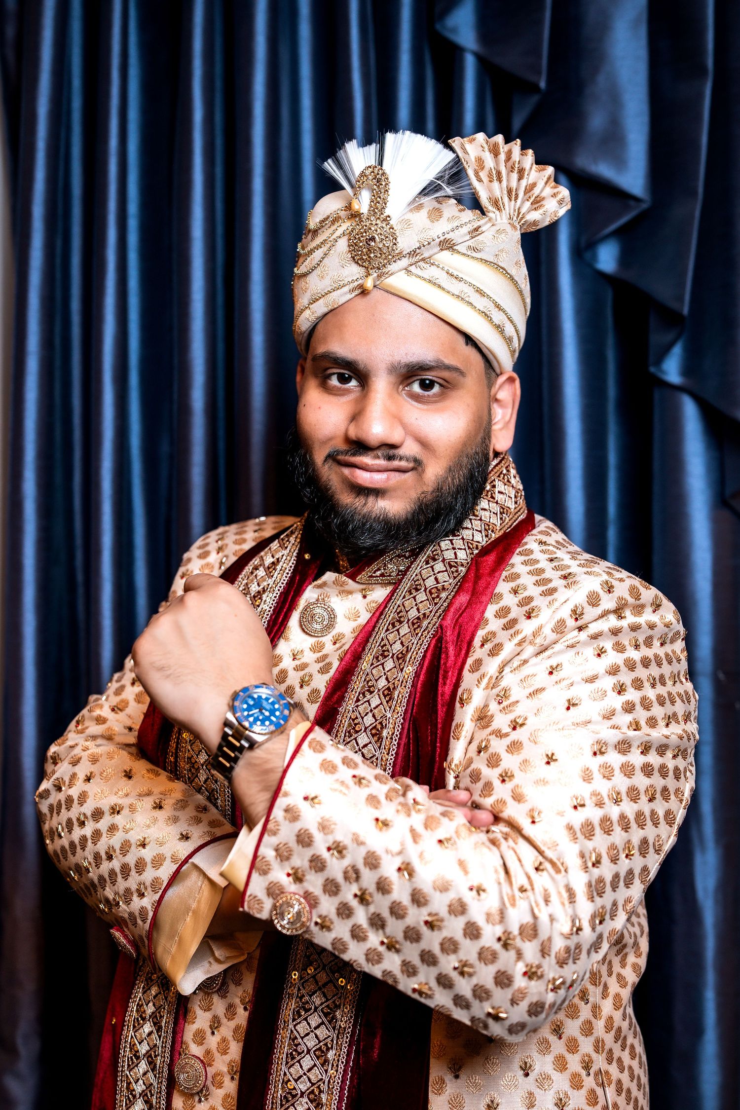 Indian groom in ornate gold sherwani and turban posing confidently against blue curtain backdrop.