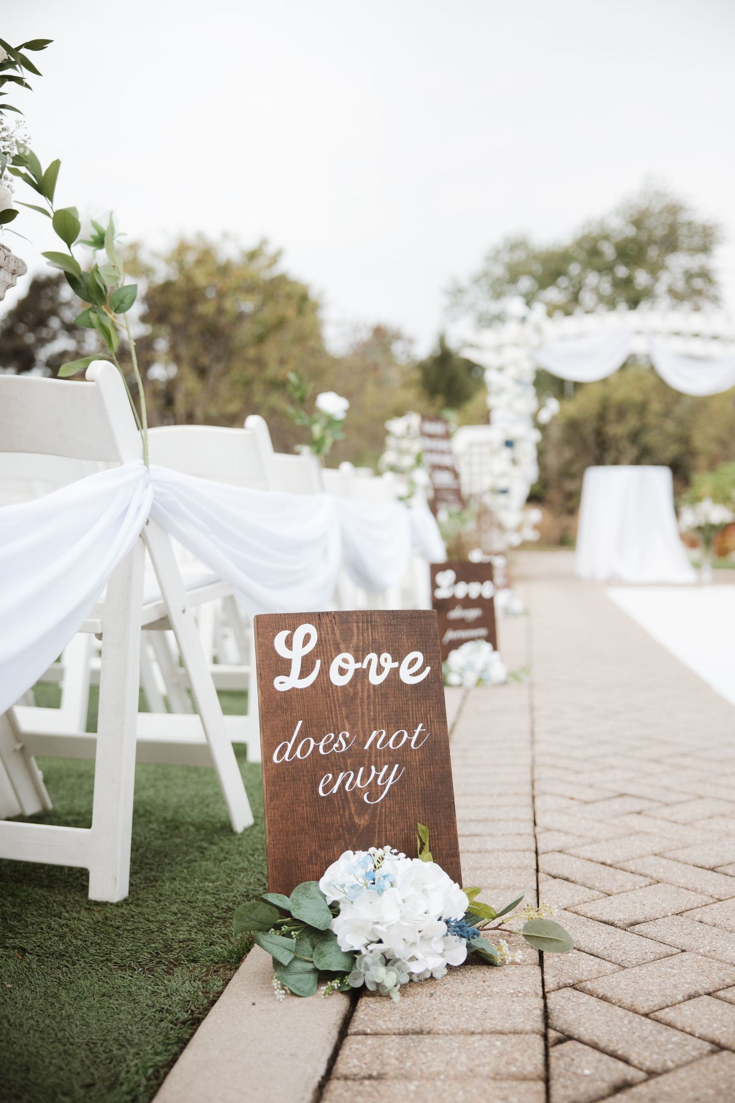 Wooden wedding sign reading Love does not envy beside white hydrangeas along a brick ceremony aisle.