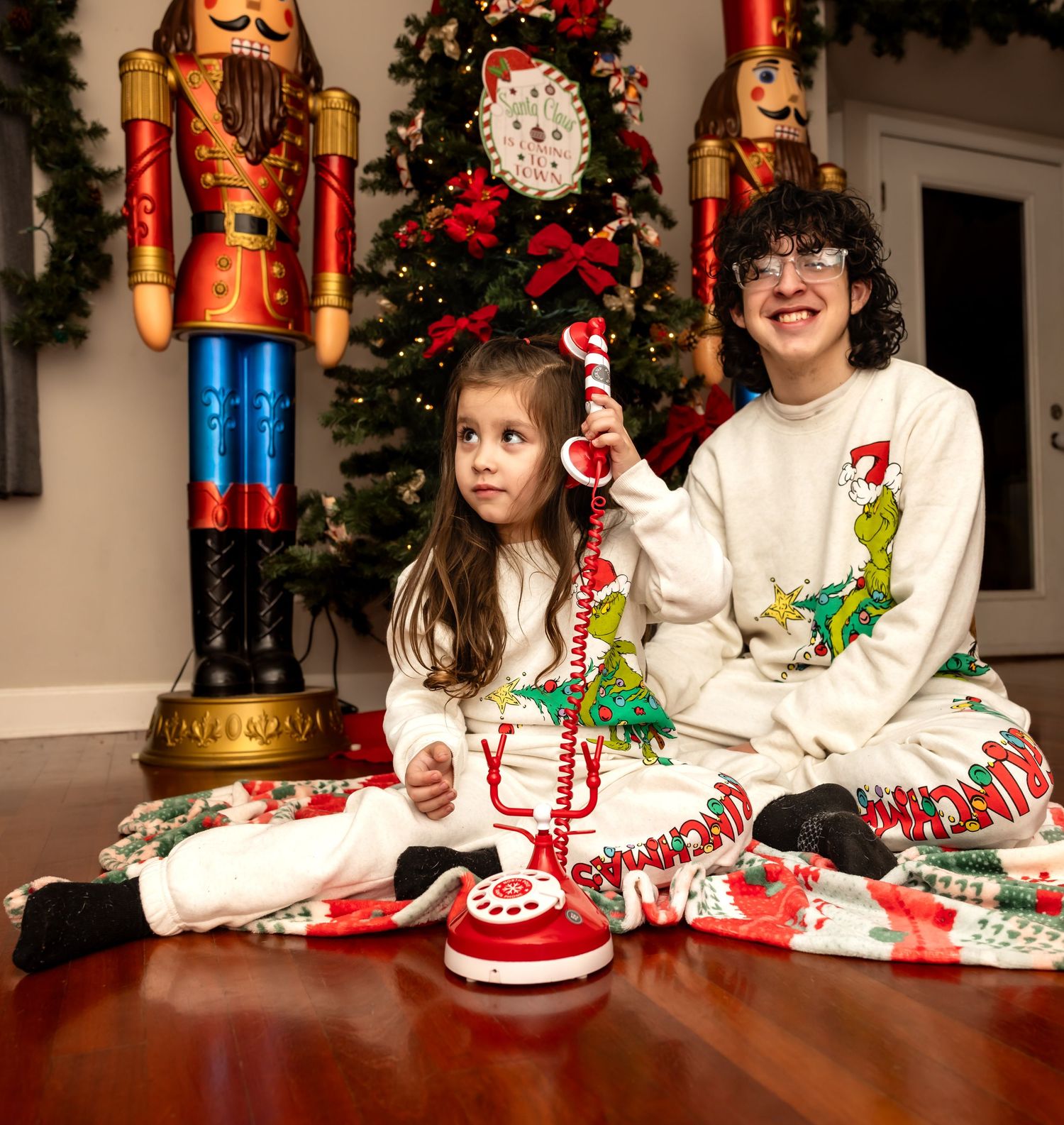 Two children in Grinch Christmas pajamas sitting on a holiday blanket with a toy phone near a decorated tree.