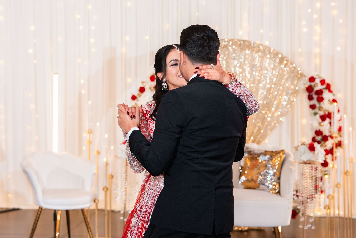 Bride in red embroidered lehenga sharing first dance with groom in black tuxedo at elegant wedding reception.