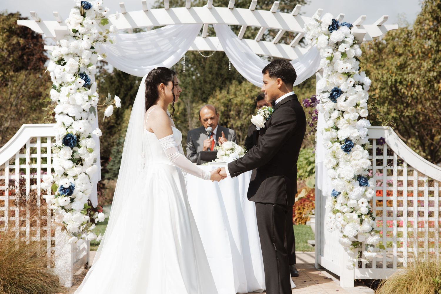 Couple holds hands before officiant under a white arch adorned with blue and white flowers.