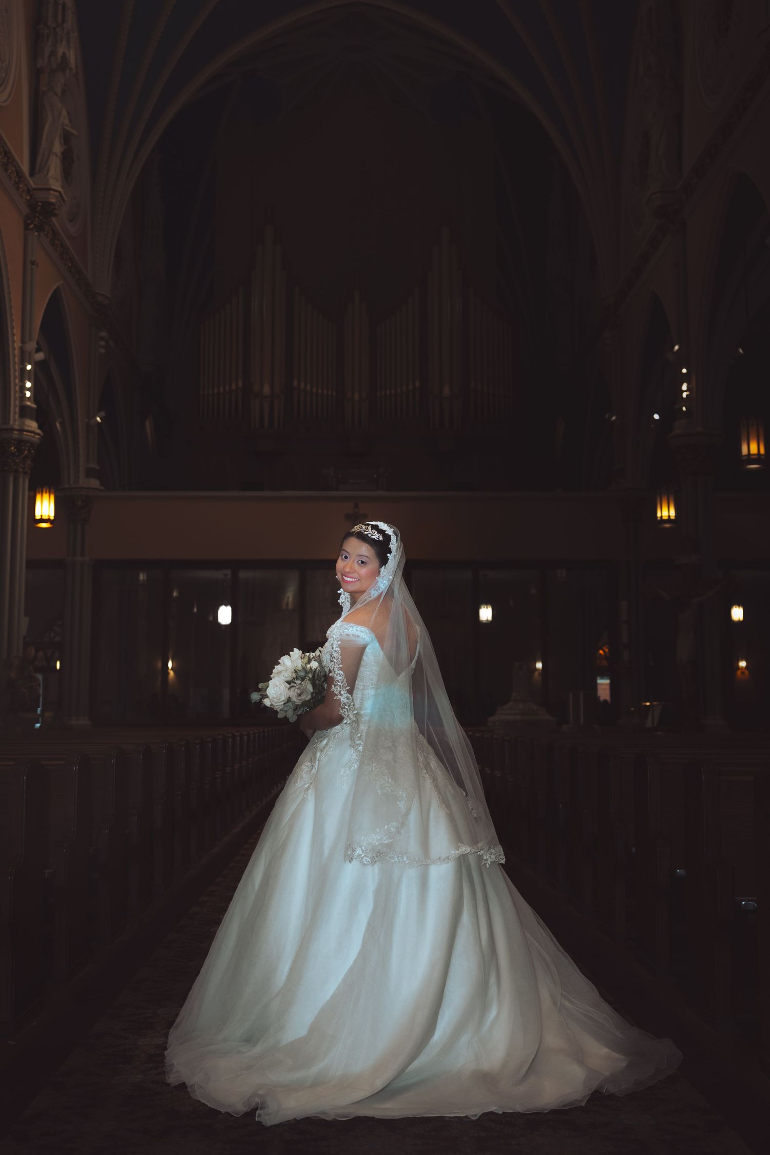 Bride in full white ball gown with lace veil stands in dimly lit church aisle with pipe organ behind.