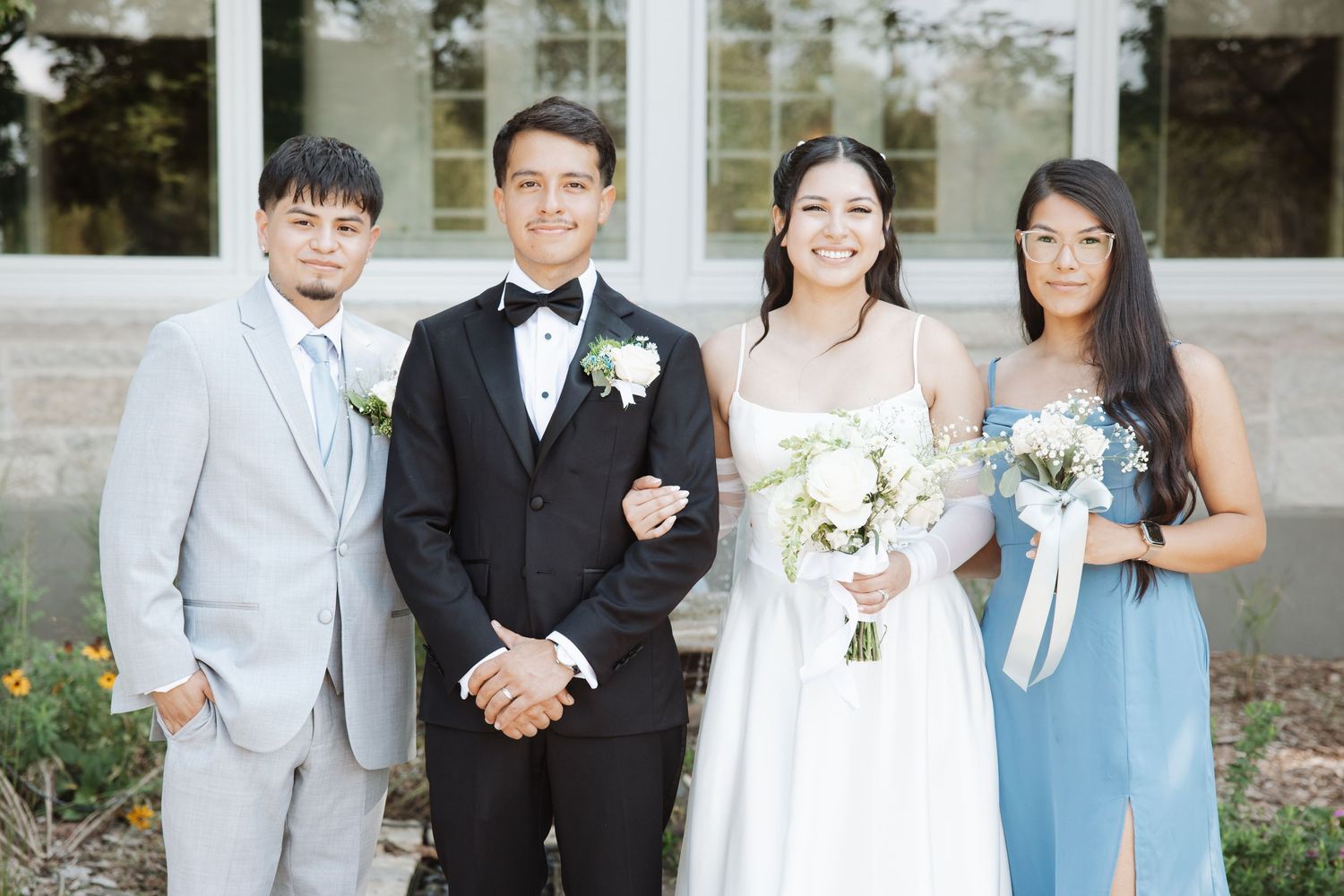 Wedding party posing outdoors with bride holding white bouquet, groom in tuxedo, groomsman in gray suit, and bridesmaid in blue dress.