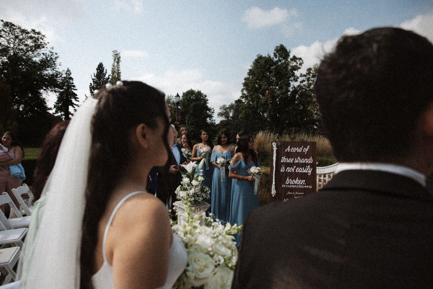 Bride in white veil walks toward bridesmaids in blue dresses at an outdoor wedding ceremony.