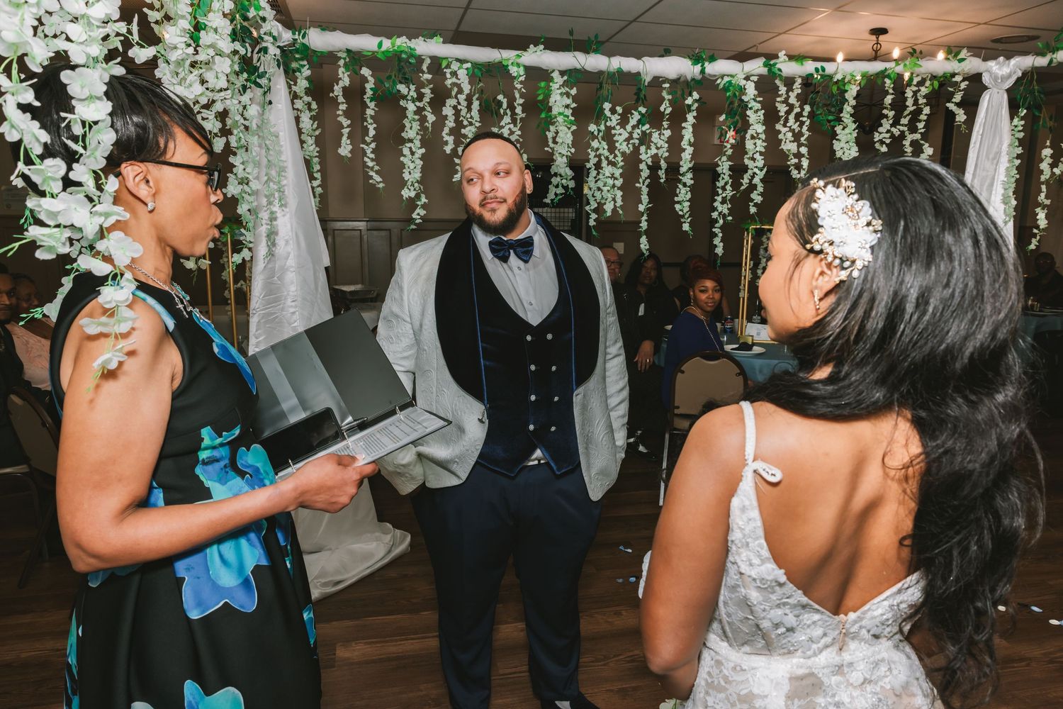 Officiant reads vows as bride and groom stand together beneath hanging white floral greenery at indoor wedding.