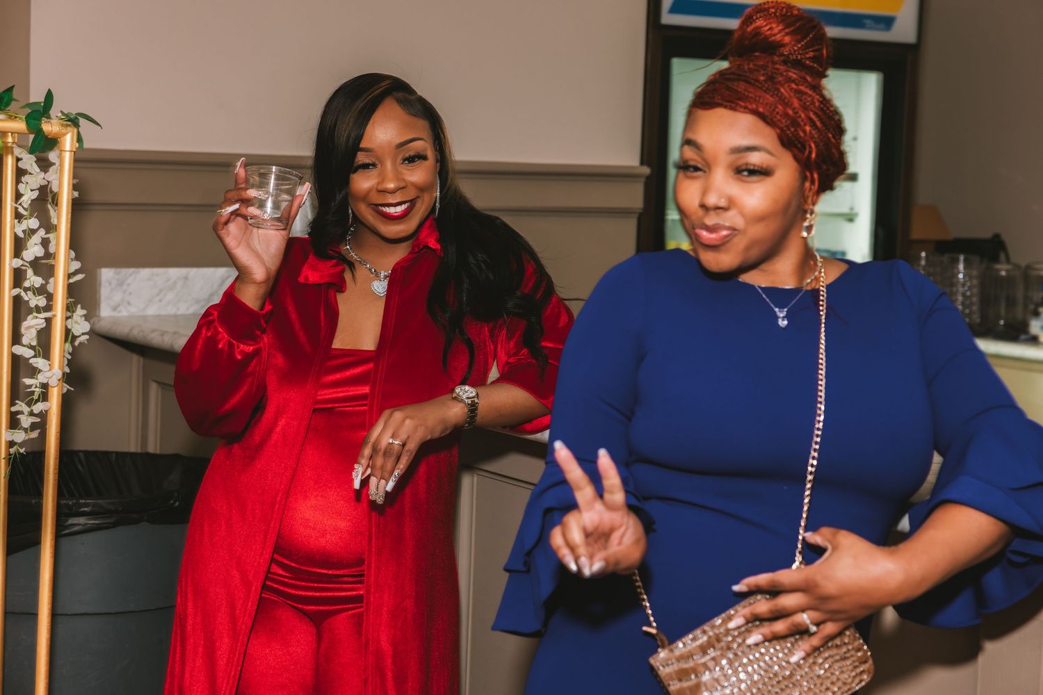 Two women smiling at a party, one in red dress holding a glass, other in blue dress making a peace sign.