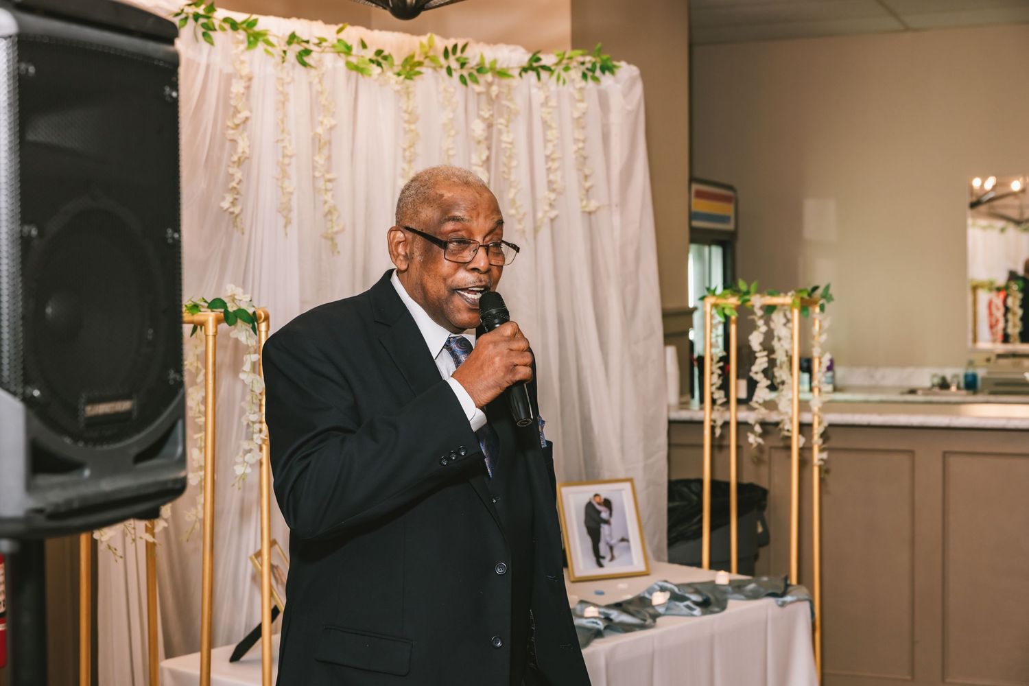 Elderly man in dark suit speaking into microphone at elegant wedding reception with floral backdrop.