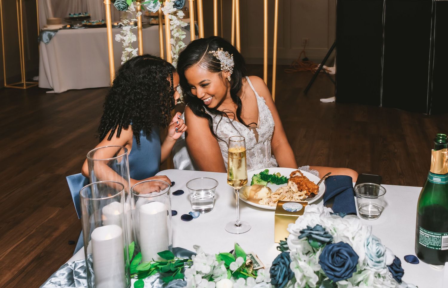 Bride in lace gown laughing with guest at wedding reception table with champagne and candles.