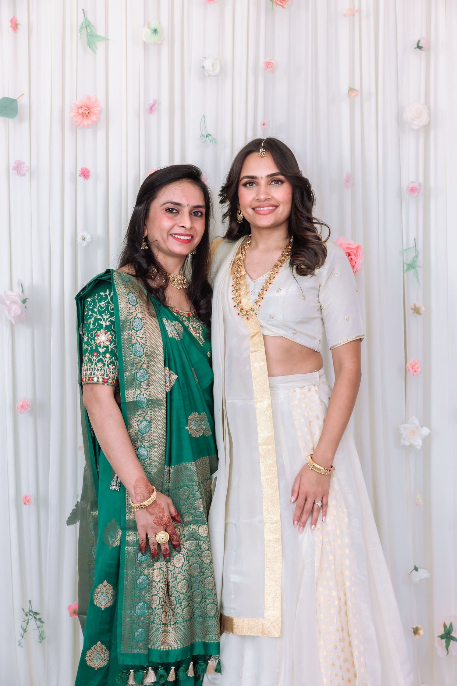 Indian mother in green saree and bride in white lehenga smile together at wedding celebration backdrop.