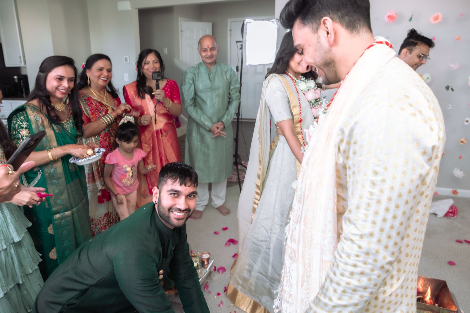 Family members smile and celebrate during traditional Indian wedding ceremony with rose petals on floor.