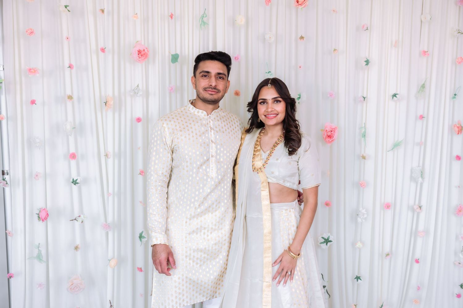 Indian couple in matching white outfits pose together at a traditional engagement celebration.