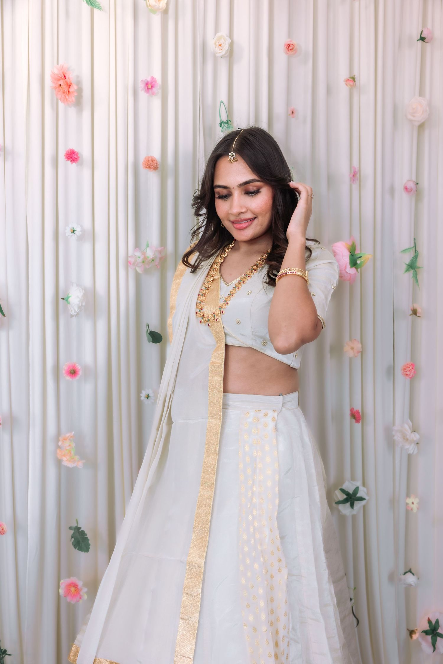 Indian woman in white lehenga choli with gold border dupatta smiling at floral backdrop.