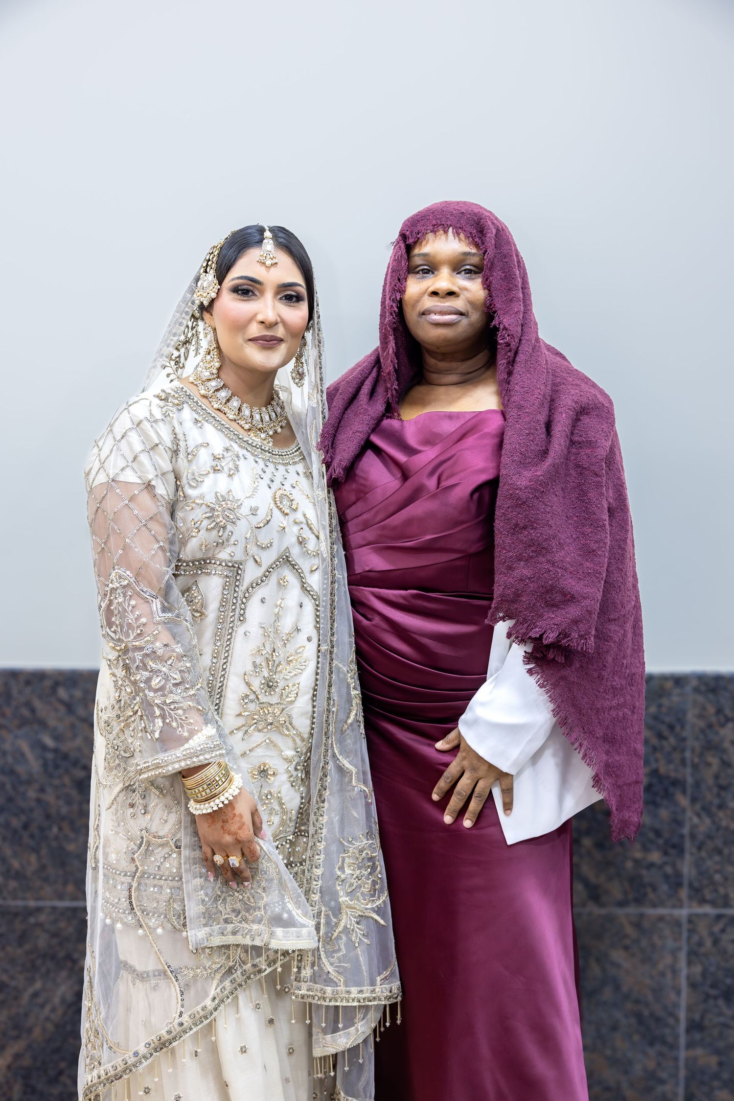 A bride in white embroidered South Asian bridal outfit poses with a woman in a burgundy dress and matching hijab.