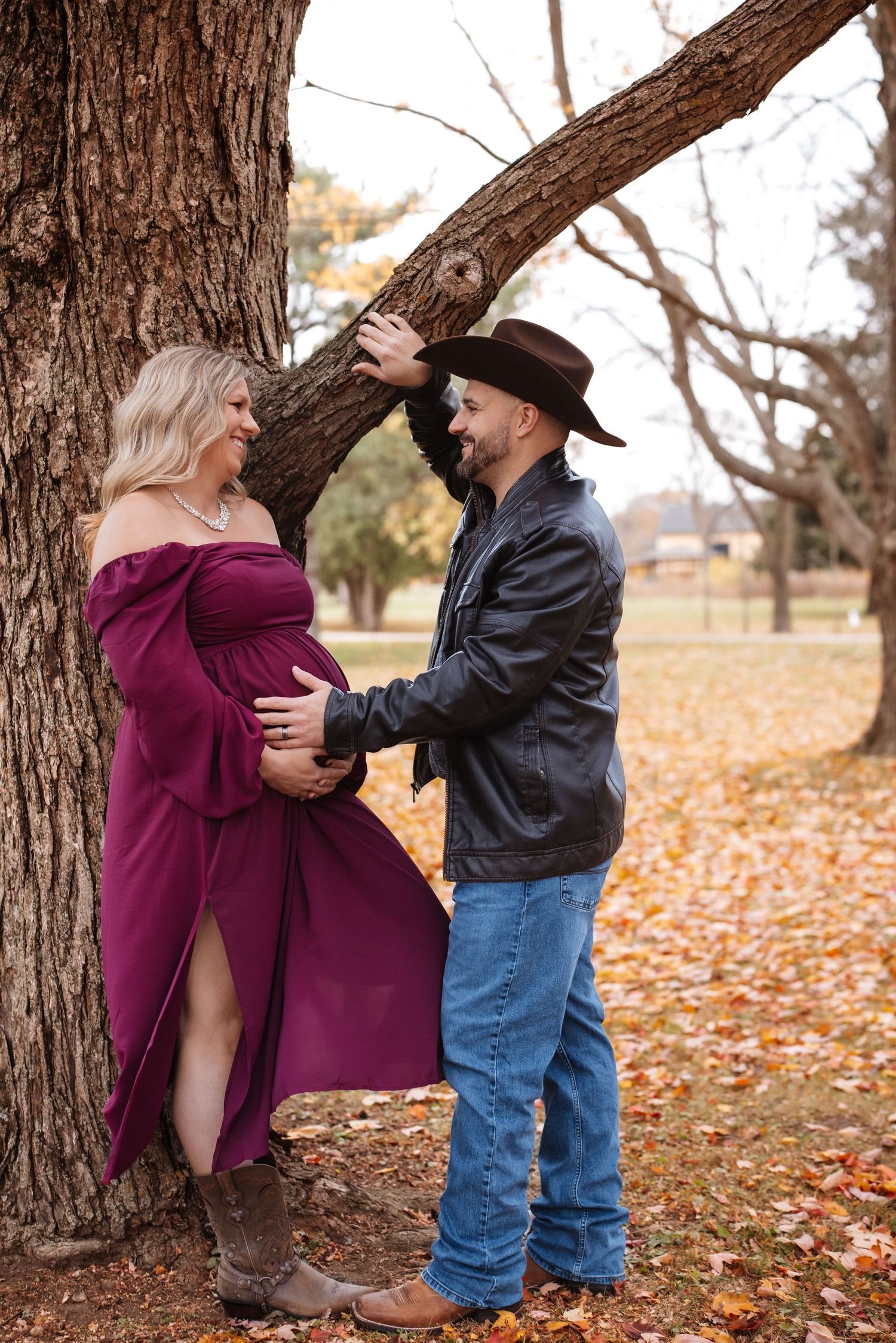 Smiling pregnant woman in off-shoulder burgundy dress with cowboy partner by tree in autumn.