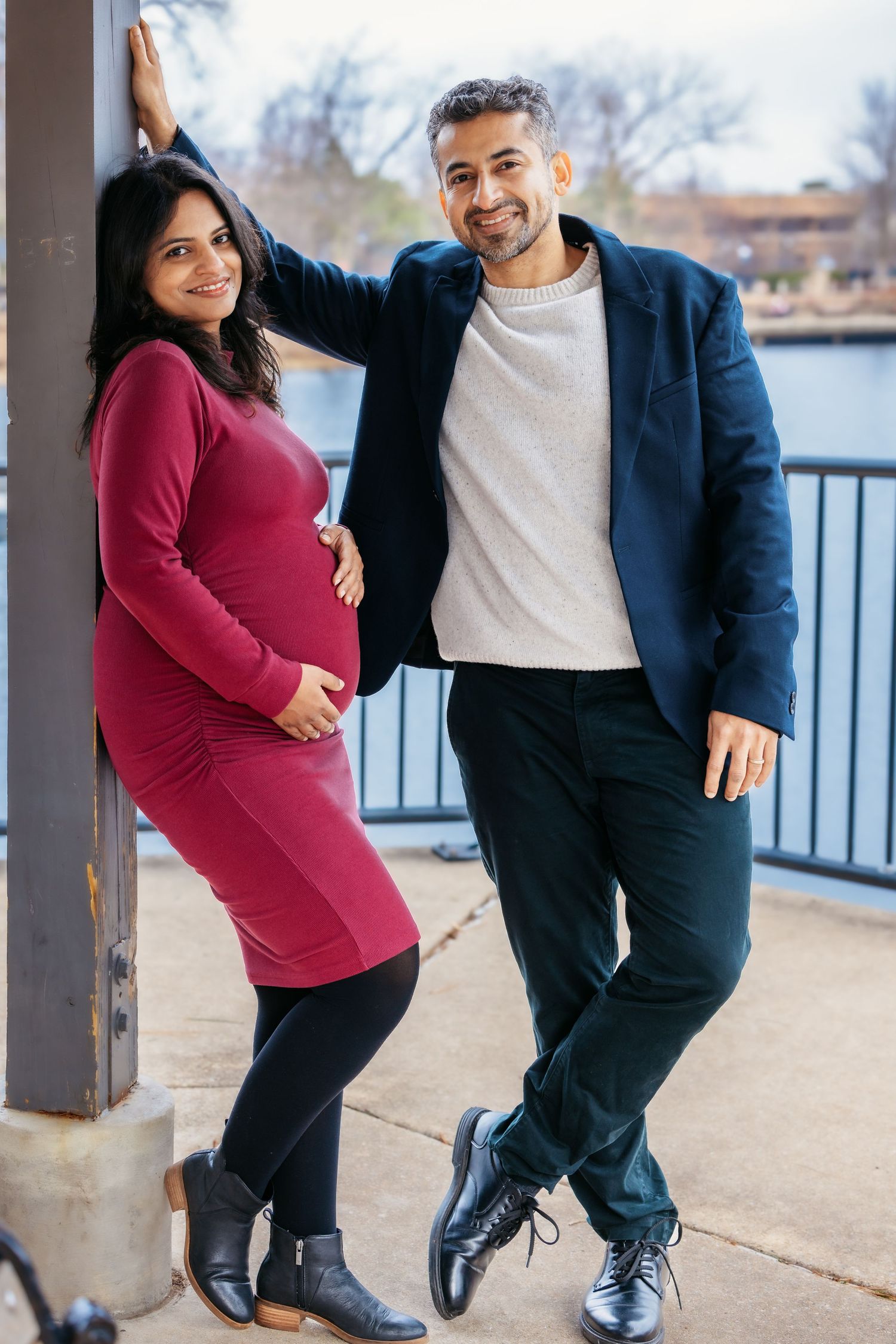 Smiling pregnant couple posing together near a waterfront bridge railing.
