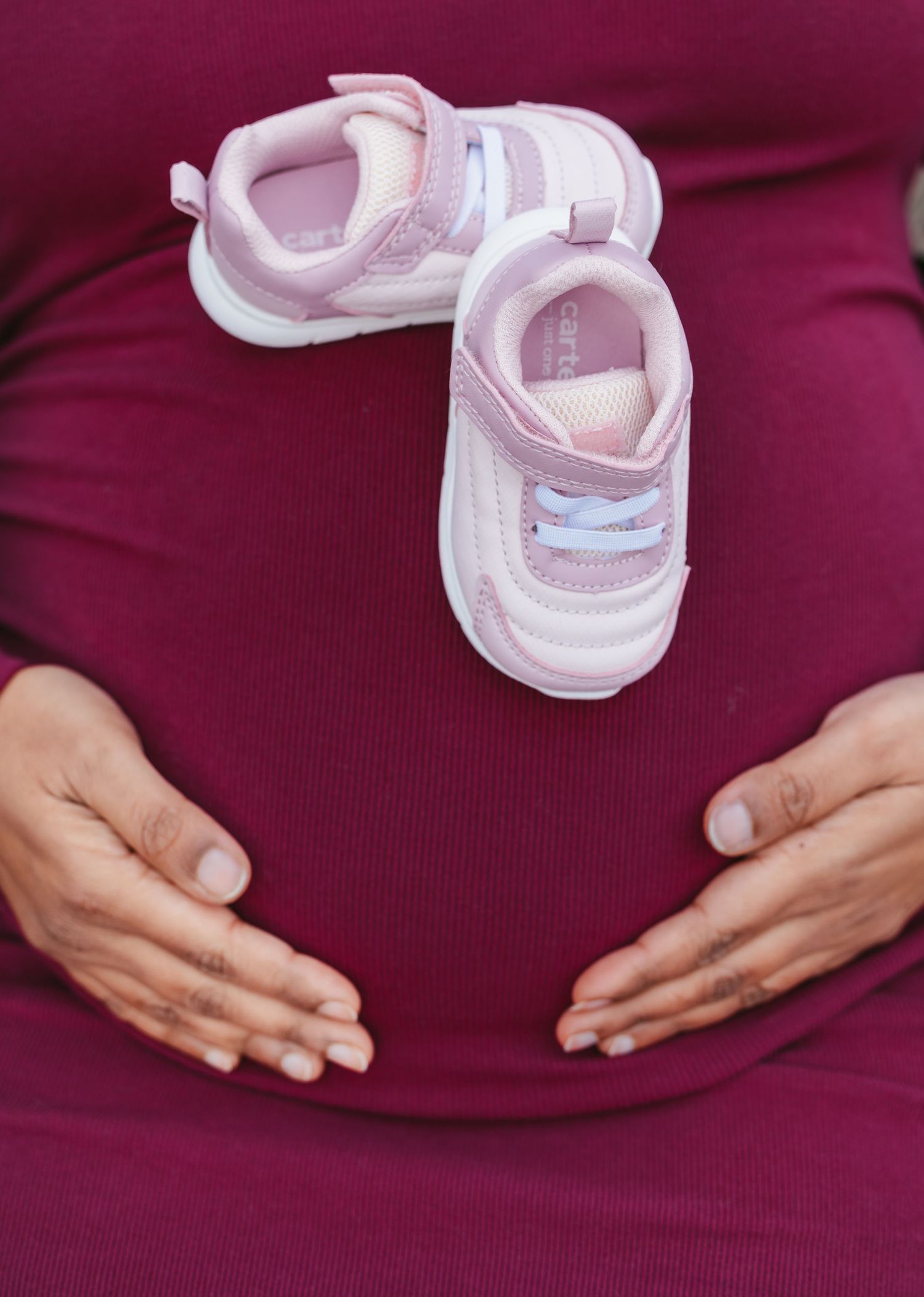 Pregnant woman in magenta dress cradling bump with small pink baby sneakers resting on top.