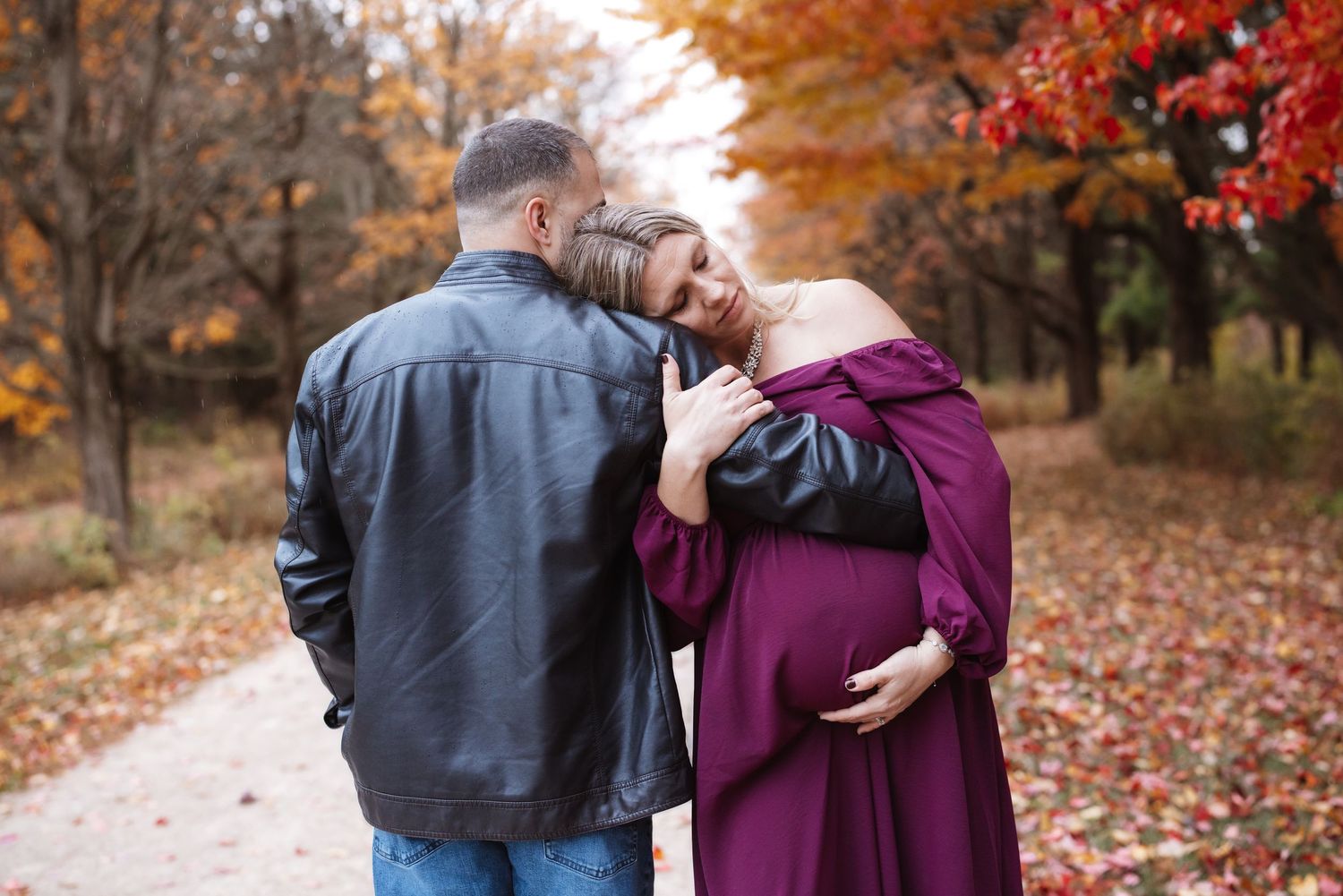 Couple sharing intimate moment on leaf-covered autumn path, pregnant woman in purple dress leans against partners shoulder.