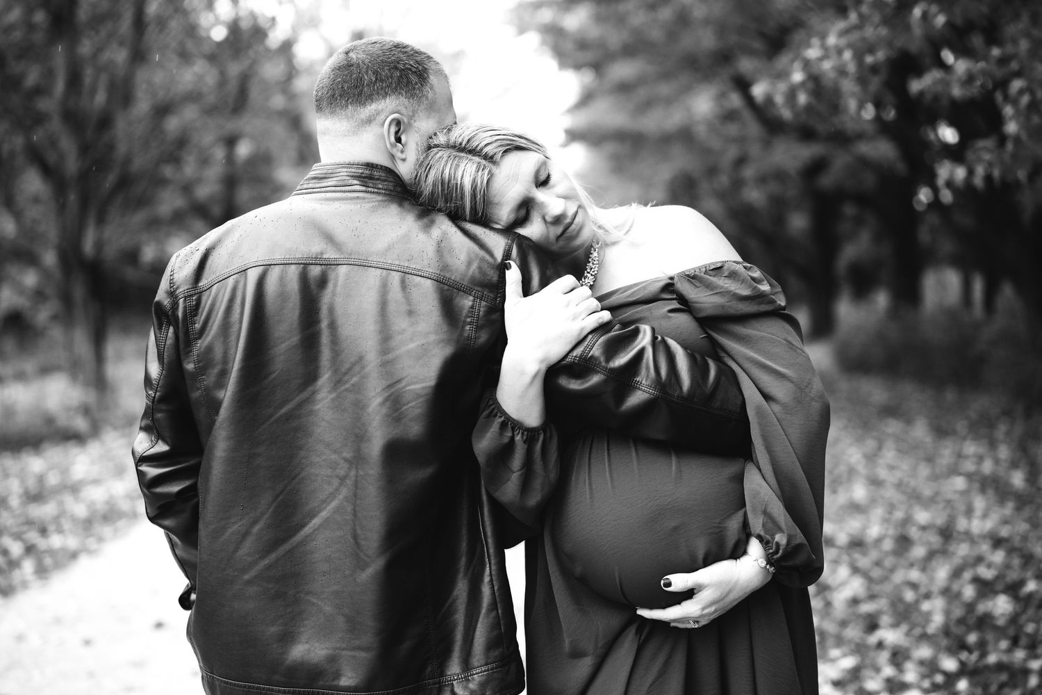 Black and white maternity photo of couple embracing on tree-lined path, woman cradling baby bump in flowing dress.