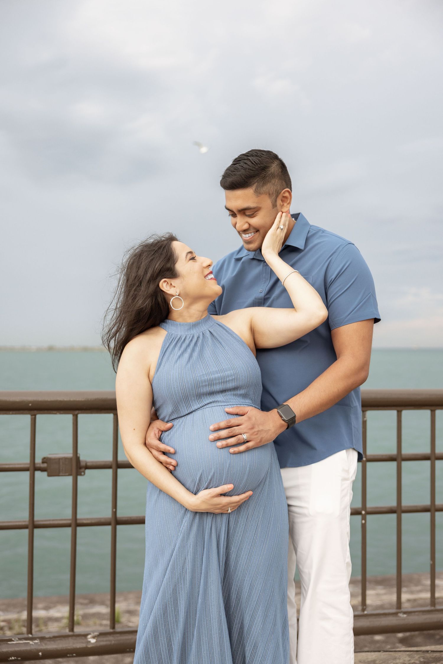 Smiling pregnant couple in blue outfits embracing tenderly by a waterfront railing under cloudy skies.