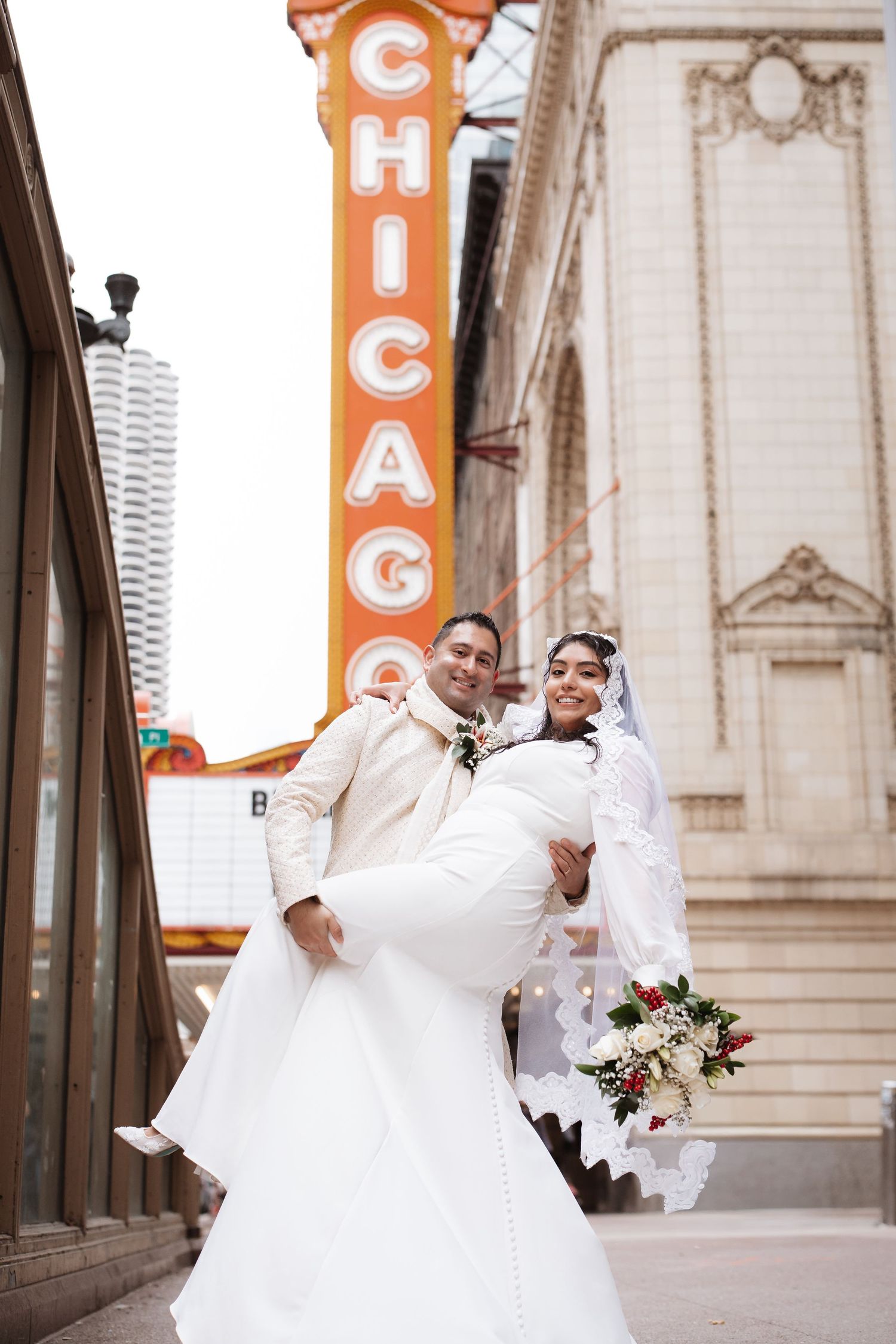 Groom lifting bride in white dress and veil outside iconic Chicago Theatre marquee sign.