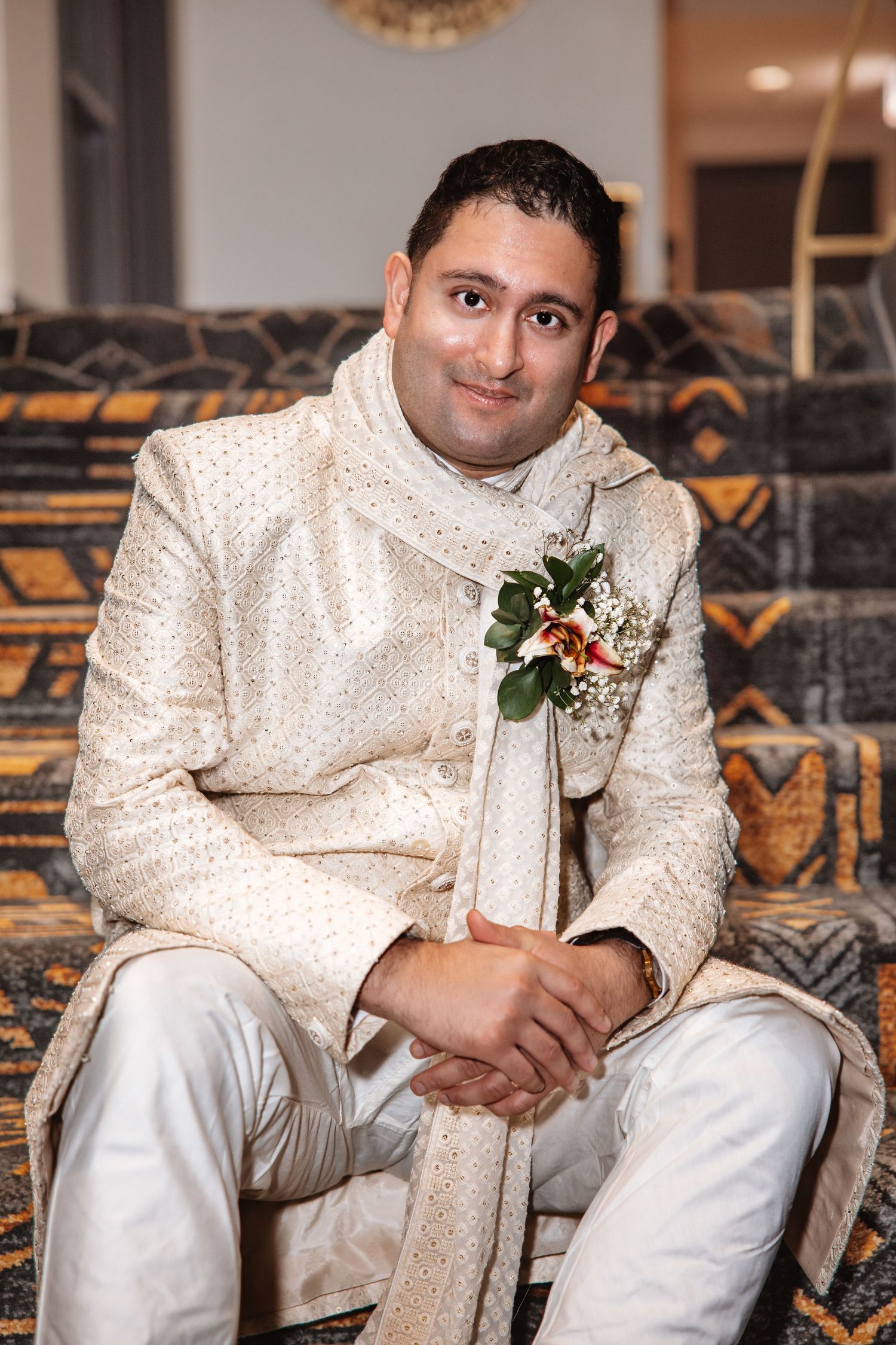 Indian groom in ivory embroidered sherwani with floral boutonniere poses seated indoors.