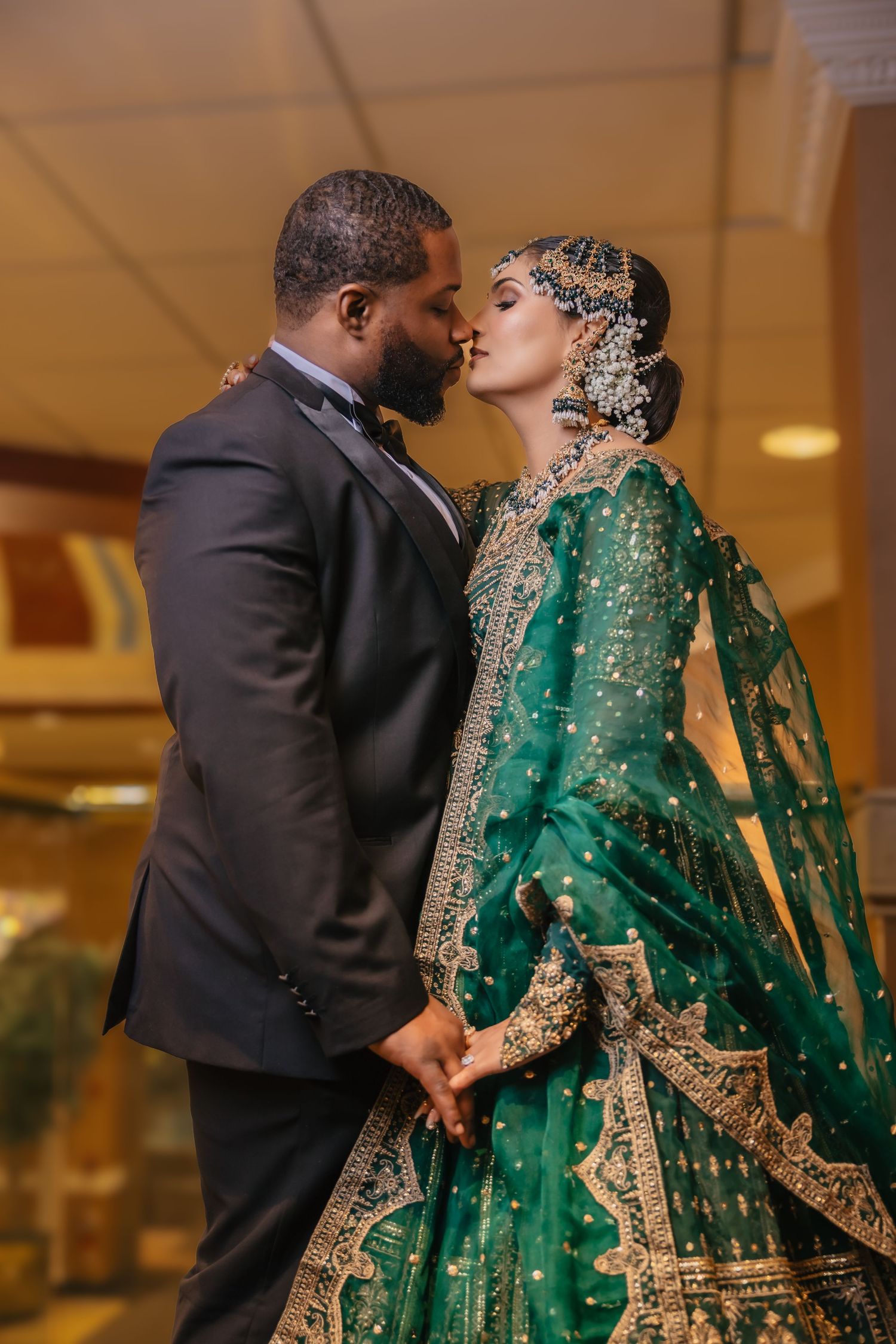 Interracial wedding couple share a kiss, bride in emerald green and gold lehenga with bridal jewelry.