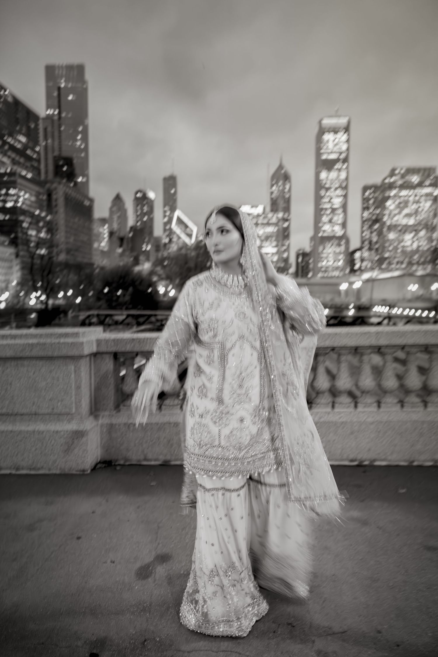 Black and white portrait of South Asian bride in ornate lehenga posing before Chicago skyline at night.