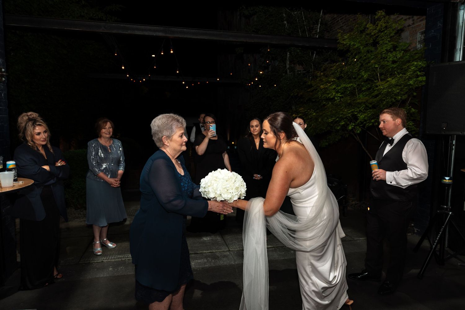 Bride in white gown receives bouquet from older woman while guests watch at outdoor wedding.